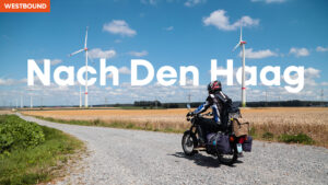Thumbnail: Westbound - Mitder Simson nach Den Haag. Man sieht eine bepackte Simson auf einer Schotterstraße. Im Hintergrund sind moderne Windkraftanlagen.